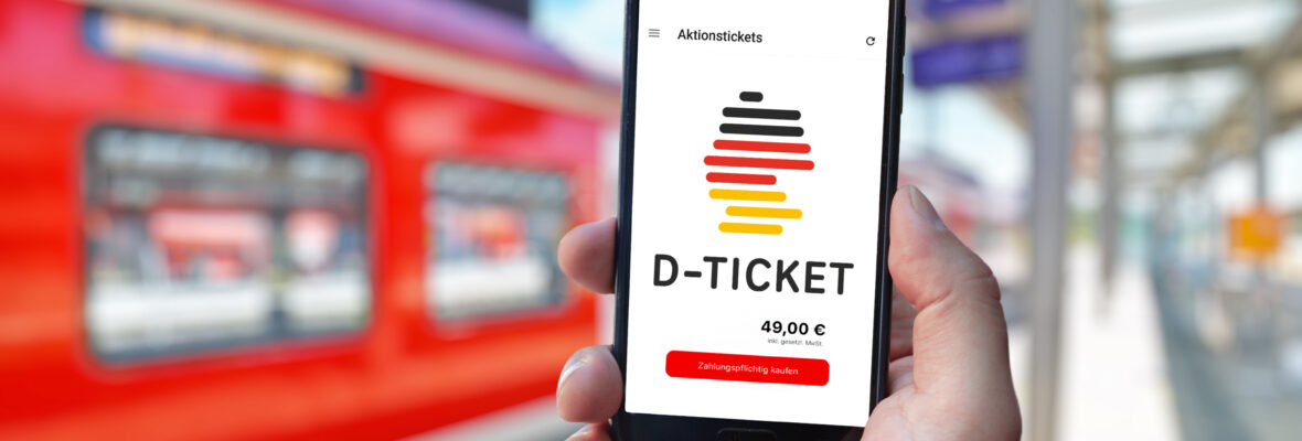 Handy in einer Hand auf dem ein Deutschlandticket zu sehen ist. Im Hintergrund fährt ein Zug auf einem Bahnsteig ein.