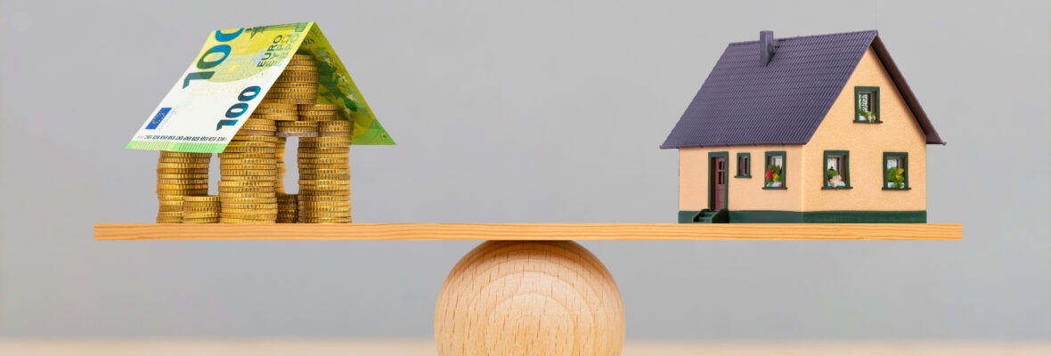 Eine kleine Holzwaage. Auf der einen Seite ein Haus aus Geld auf der anderen ein Modelhaus.