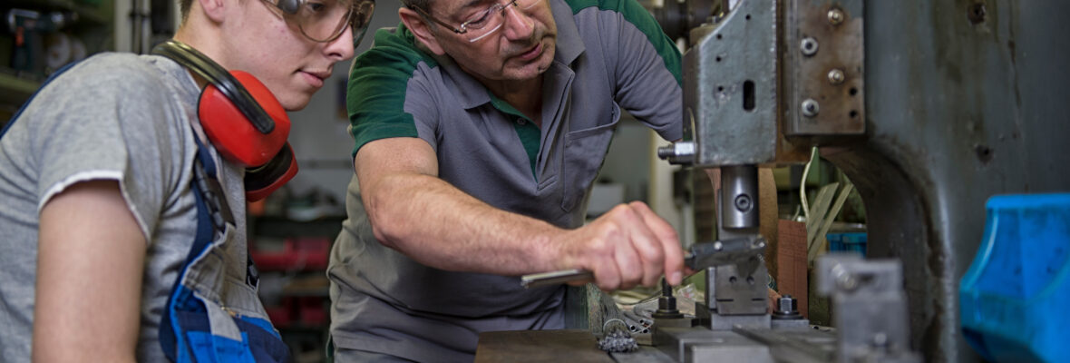 Ein älterer Meister erklärt einem Schüler das Bohren mit einer Standbohrmaschine.